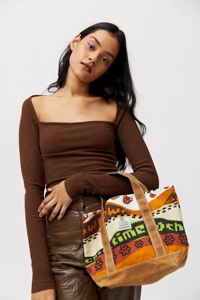May include: Brown tote bag with a colorful patterned design, featuring orange, green, and white elements. The bag has brown handles and a brown base. The model is wearing a brown long-sleeve top.