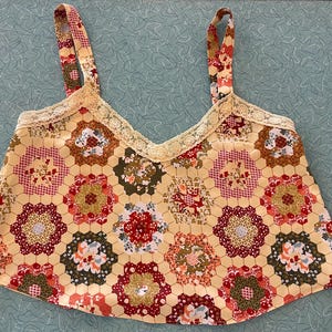 May include: A yellow patchwork top with floral and geometric patterns. The top features a V-neckline trimmed with lace and matching patterned straps. The patchwork design includes red, brown, and green floral motifs.