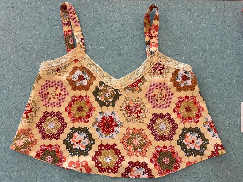 May include: A yellow patchwork top with floral and geometric patterns. The top features a V-neckline trimmed with lace and matching patterned straps. The patchwork design includes red, brown, and green floral motifs.