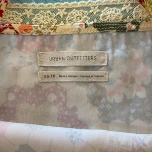 May include: Close-up of a floral patterned top with the "Urban Outfitters" label. The top features a mix of colors, including white, pink, and green, with a delicate lace trim. The size tag reads "XS-TP Made in Vietnam".