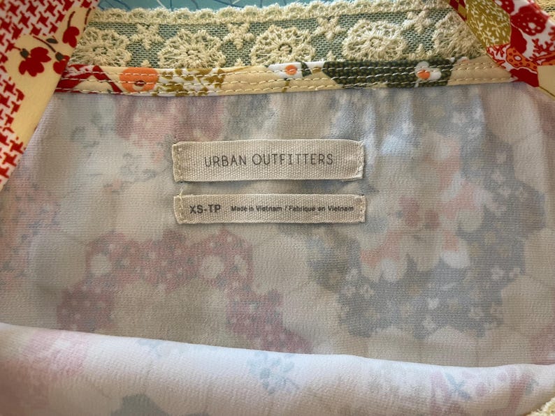 May include: Close-up of a floral patterned top with the "Urban Outfitters" label. The top features a mix of colors, including white, pink, and green, with a delicate lace trim. The size tag reads "XS-TP Made in Vietnam".