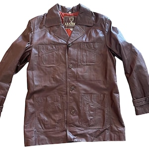 May include: A brown leather jacket with a button-up front and multiple pockets. The jacket has a label that reads "Leather of Distinction".