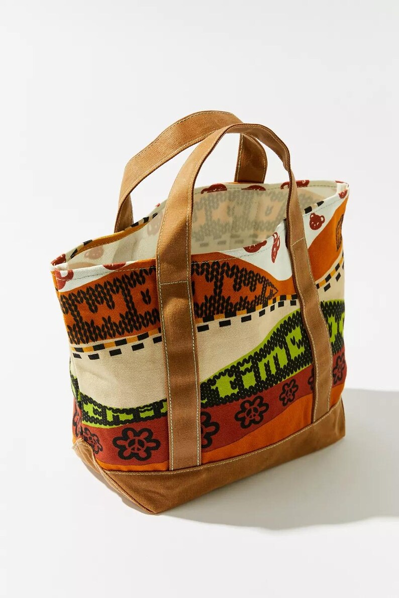 May include: A canvas tote bag with a colorful patterned design. The bag features brown handles and a brown base. The pattern includes orange, green, and cream colors with floral and geometric shapes.