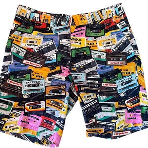 May include: Black shorts covered in a colorful pattern of cassette tapes. The tapes are in various colors, including yellow, green, pink, and blue, with text labels. A fun and retro design.