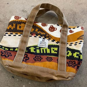 May include: A colorful tote bag with a brown corduroy base and handles. The bag features a repeating pattern of brown bear faces, floral designs, and geometric shapes in shades of brown, orange, green, and cream. A small tag is visible on the front.