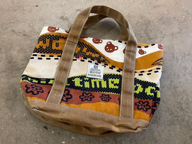May include: A colorful tote bag with a brown corduroy base and handles. The bag features a repeating pattern of brown bear faces, floral designs, and geometric shapes in shades of brown, orange, green, and cream. A small tag is visible on the front.