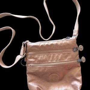 May include: A rose gold crossbody bag with a zipper closure and a Kipling logo. The bag has a long adjustable strap.