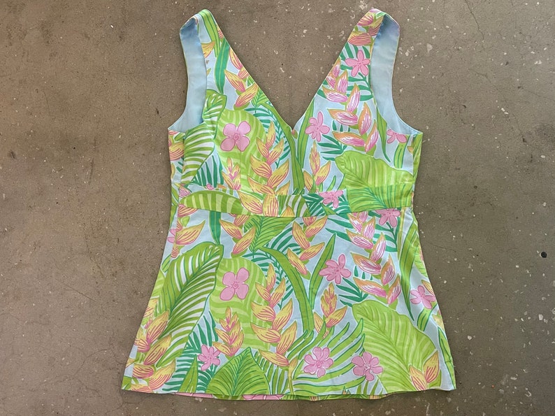 May include: A sleeveless, light blue top with a V-neckline and a tropical print featuring green leaves, pink flowers, and yellow blossoms.