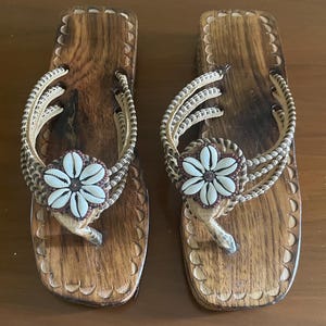 May include: A pair of wooden sandals with a natural finish. The sandals feature woven straps and a decorative flower made of seashells. The footbed is carved with a scalloped edge, and the thong is made of a light brown material.