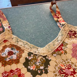 May include: Close-up of a patchwork top with floral and geometric patterns in red, green, and cream. The top features a cream-colored lace trim along the neckline and straps. The straps are patterned with a mix of floral and geometric designs.