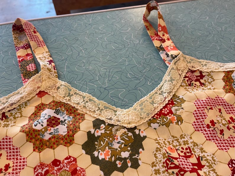 May include: Close-up of a patchwork top with floral and geometric patterns in red, green, and cream. The top features a cream-colored lace trim along the neckline and straps. The straps are patterned with a mix of floral and geometric designs.