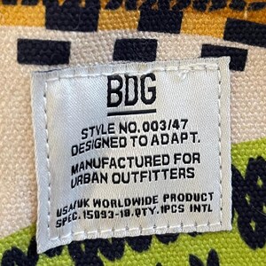 May include: A white tag with black text reading "BDG STYLE NO.003/47 DESIGNED TO ADAPT. MANUFACTURED FOR URBAN OUTFITTERS USA/UK WORLDWIDE PRODUCT SPEC. 15093-18.QTY.1PCS INTL." The tag is on a patterned fabric.