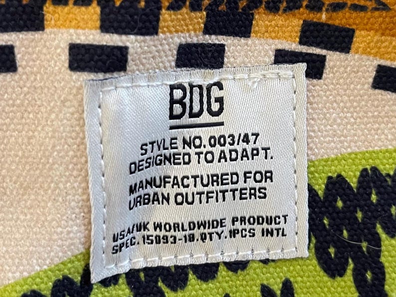 May include: A white tag with black text reading "BDG STYLE NO.003/47 DESIGNED TO ADAPT. MANUFACTURED FOR URBAN OUTFITTERS USA/UK WORLDWIDE PRODUCT SPEC. 15093-18.QTY.1PCS INTL." The tag is on a patterned fabric.