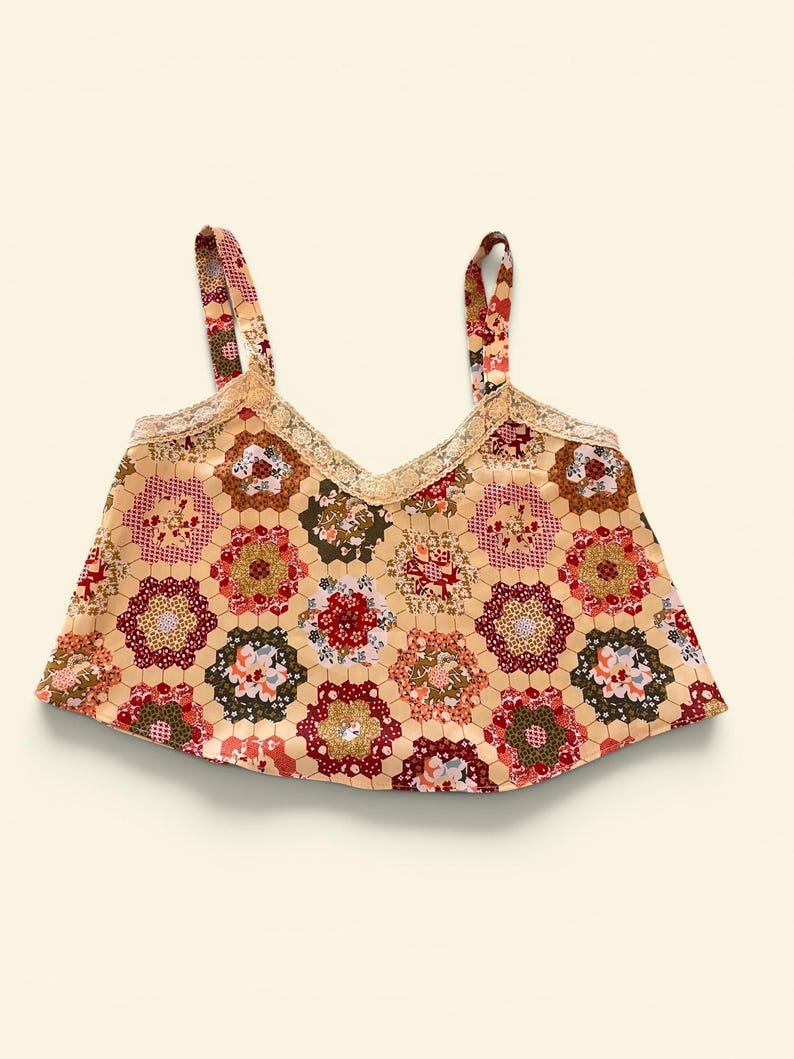 May include: A beige crop top with a floral patchwork design. The top features a V-neckline trimmed with lace and thin straps. The floral pattern includes red, green, and brown accents.