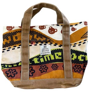 May include: A canvas tote bag with brown handles and a colorful patterned design. The pattern includes brown bear cup illustrations, floral and geometric shapes, and the text "BDG". The bag has a rectangular shape and a sturdy base.