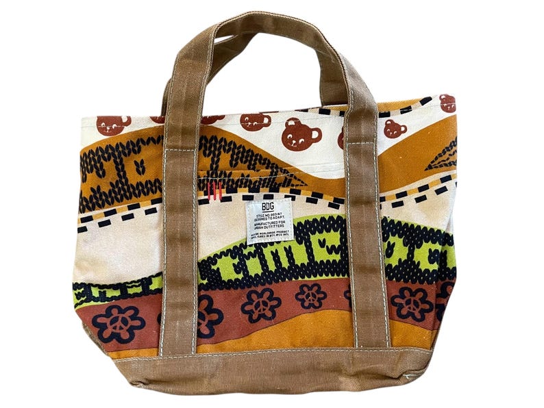 May include: A canvas tote bag with brown handles and a colorful patterned design. The pattern includes brown bear cup illustrations, floral and geometric shapes, and the text "BDG". The bag has a rectangular shape and a sturdy base.