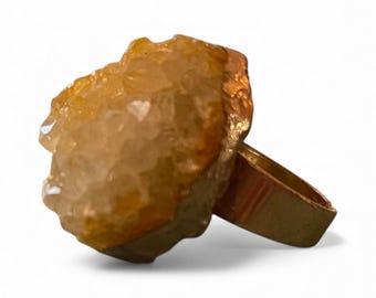 May include: A gold-toned ring featuring a large, rough-cut citrine gemstone. The citrine has a warm, amber color with a textured surface. The ring's band is a simple, polished gold metal.