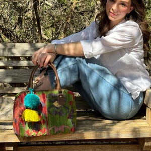May include: A woman wearing a white shirt and blue jeans sits on a wooden bench with a colorful woven handbag with tassels. The handbag has a brown leather handle and a green, red, and yellow pattern.