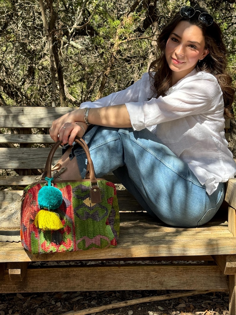 May include: A woman wearing a white shirt and blue jeans sits on a wooden bench with a colorful woven handbag with tassels. The handbag has a brown leather handle and a green, red, and yellow pattern.
