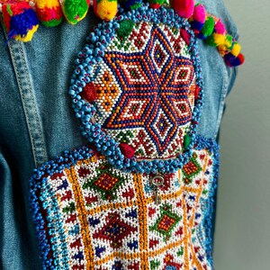 Pode incluir: Uma jaqueta de ganga com bordado de contas coloridas nas costas. O bordado apresenta um desenho de estrela com padr&otilde;es geom&eacute;tricos em tons de azul, vermelho, laranja, verde e branco. A jaqueta tamb&eacute;m tem uma fileira de pompons coloridos ao longo da borda superior.