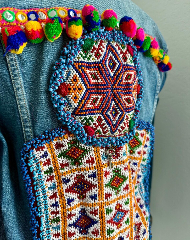 Pode incluir: Uma jaqueta de ganga com bordado de contas coloridas nas costas. O bordado apresenta um desenho de estrela com padr&otilde;es geom&eacute;tricos em tons de azul, vermelho, laranja, verde e branco. A jaqueta tamb&eacute;m tem uma fileira de pompons coloridos ao longo da borda superior.