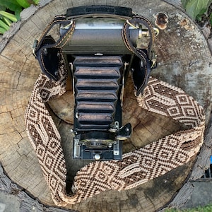 May include: A vintage black camera with a brown and white patterned strap. The camera has a bellows system and a lens with a shutter.
