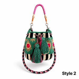 May include: A bucket bag featuring a woven pattern of black and cream stripes with green, pink, and tan geometric accents. The bag includes a pink braided handle, a long multi-colored shoulder strap, and two large green tassels. Text reads Style 2.