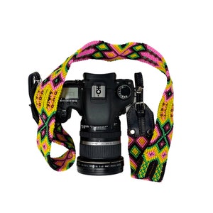 May include: A black DSLR camera with a colorful woven strap. The strap features a geometric pattern in pink, yellow, green, and black. The camera lens is black with white text.