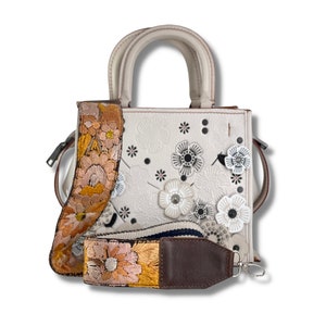 Puede incluir: Bolso de mano de cuero blanco con un diseño floral y una correa con estampado floral marrón. El bolso tiene un asa superior y una correa para el hombro desmontable.