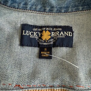 Pode incluir: Um r&oacute;tulo de jaqueta jeans Lucky Brand preto e dourado com o texto "The Art of Blue Jeans" e "Lucky Brand" em fonte dourada. O r&oacute;tulo tamb&eacute;m inclui o texto "Registered", "Trademark" e "Too Tough to Die". O tamanho "S" e "Made in China" s&atilde;o impressos em um r&oacute;tulo branco menor anexado ao r&oacute;tulo principal.
