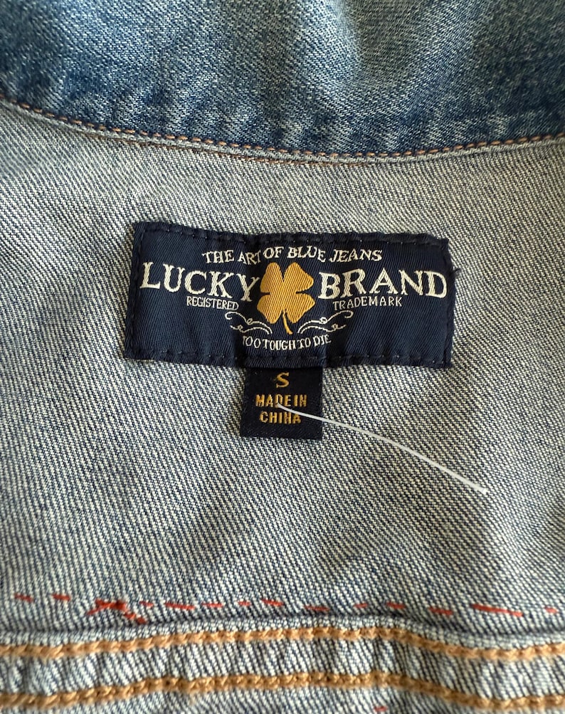 Pode incluir: Um r&oacute;tulo de jaqueta jeans Lucky Brand preto e dourado com o texto "The Art of Blue Jeans" e "Lucky Brand" em fonte dourada. O r&oacute;tulo tamb&eacute;m inclui o texto "Registered", "Trademark" e "Too Tough to Die". O tamanho "S" e "Made in China" s&atilde;o impressos em um r&oacute;tulo branco menor anexado ao r&oacute;tulo principal.