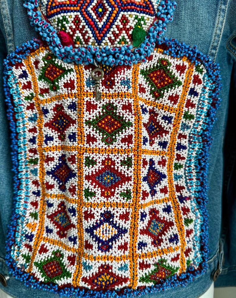 Pode incluir: Uma jaqueta de ganga com um design de contas coloridas nas costas. O design apresenta um padr&atilde;o geom&eacute;trico com quadrados e diamantes em tons de vermelho, azul, verde, branco e laranja.