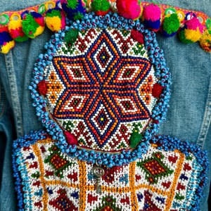 Pode incluir: Uma jaqueta de ganga com enfeites de contas coloridas. A jaqueta apresenta um grande desenho circular com um padr&atilde;o em forma de estrela no centro, rodeado por uma borda de contas azuis. A parte inferior da jaqueta tem um desenho retangular menor com um padr&atilde;o geom&eacute;trico.