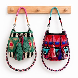 May include: Two bucket bags with woven patterns and tassels. The left bag features green, black, and white stripes with pink accents and a pink handle. The right bag displays red, blue, and black geometric patterns with a bright blue handle.