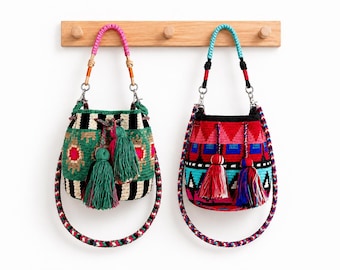 May include: Two bucket bags with woven patterns and tassels. The left bag features green, black, and white stripes with pink accents and a pink handle. The right bag displays red, blue, and black geometric patterns with a bright blue handle.