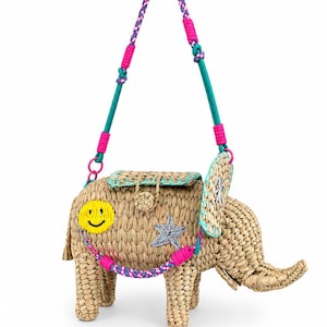 May include: A woven straw elephant-shaped bag featuring a yellow smiley face patch, a silver star appliqué, and a turquoise trim. The bag includes a multi-colored braided shoulder strap with bright pink accents and a flap closure on the back.