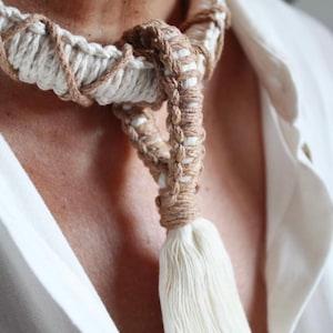 May include: A white and brown macrame necklace with a long white tassel. The necklace is made of natural fibers and has a bohemian style.