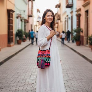 May include: A white long-sleeved dress with eyelet embroidery at the hem and a colorful crocheted crossbody bag. The bag features a geometric pattern in neon pink, orange, teal, and black, finished with a large, multi-colored yarn tassel.