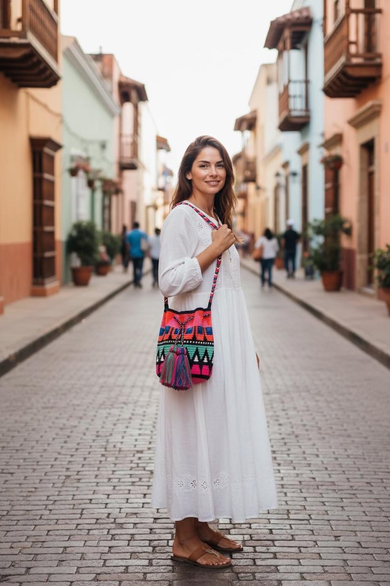 May include: A white long-sleeved dress with eyelet embroidery at the hem and a colorful crocheted crossbody bag. The bag features a geometric pattern in neon pink, orange, teal, and black, finished with a large, multi-colored yarn tassel.
