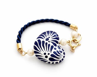 Pode incluir: Uma pulseira azul marinho e branca em forma de coração. O pingente em forma de coração apresenta um design floral azul. A pulseira tem uma faixa azul marinho em forma de corda, detalhes dourados e um fecho com duas pérolas.