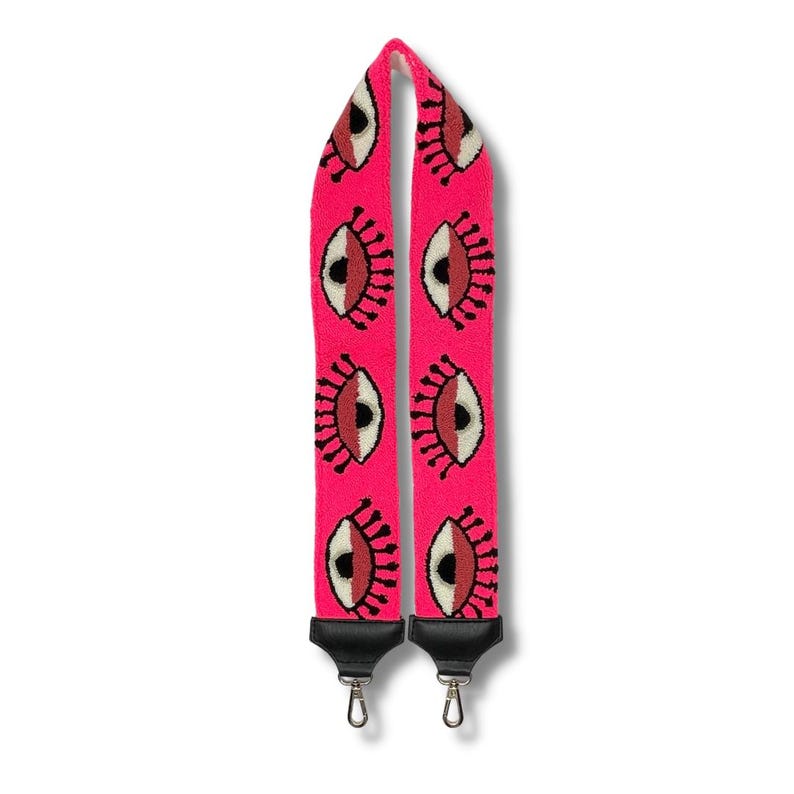 May include: A pink adjustable camera strap with a repeating pattern of black outlined eyes with red and white details.