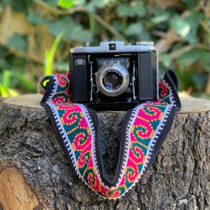 May include: A vintage silver and black camera with a black and white patterned strap. The camera has a lens with the text "Meyer-Optic Anastigmat 1:4.5 f=75mm" printed on it.