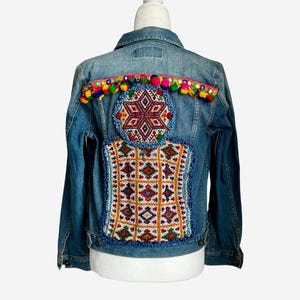 Pode incluir: Uma jaqueta de ganga azul com bordados coloridos e pompons nas costas.