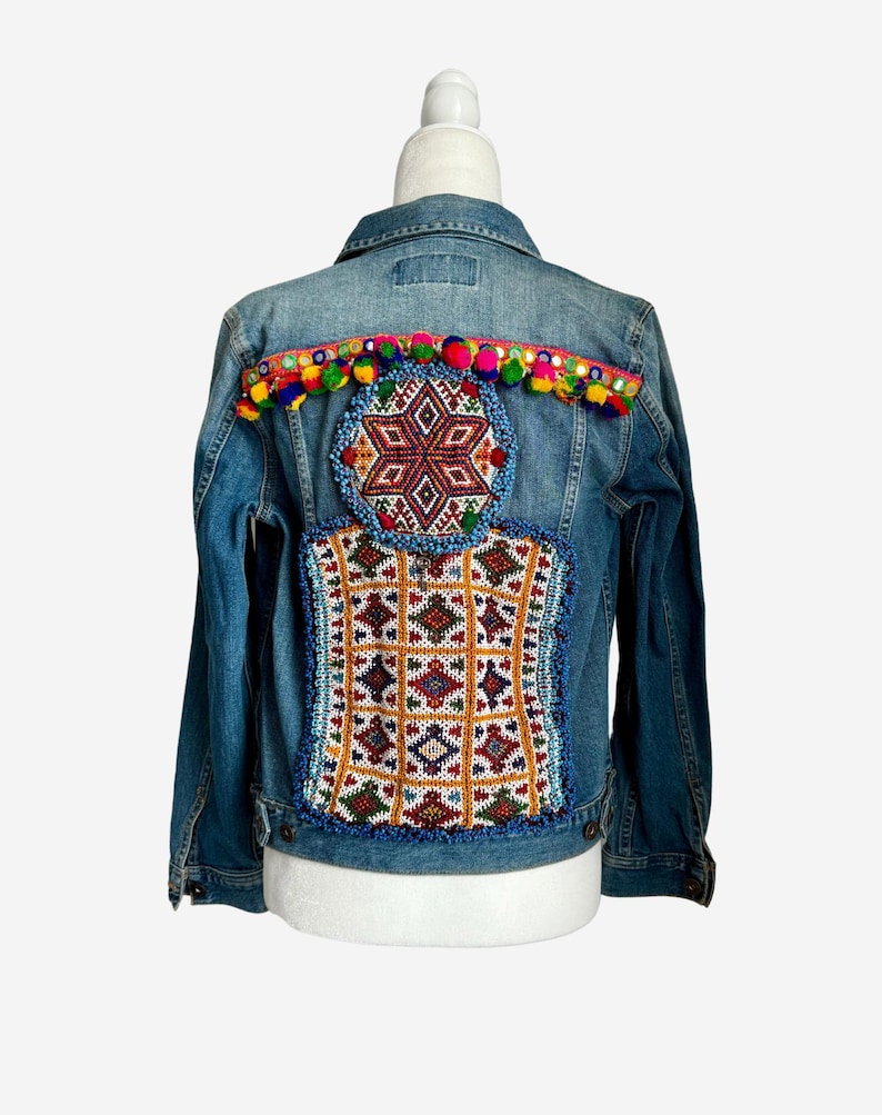 Pode incluir: Uma jaqueta de ganga azul com bordados coloridos e pompons nas costas.
