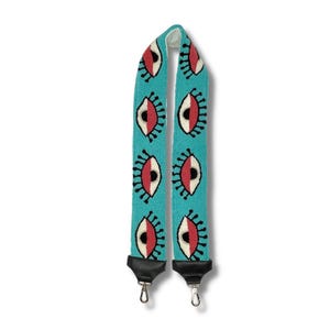 May include: A turquoise blue adjustable strap with a repeating pattern of black outlined eyes with white and red details. The strap has black metal hardware.