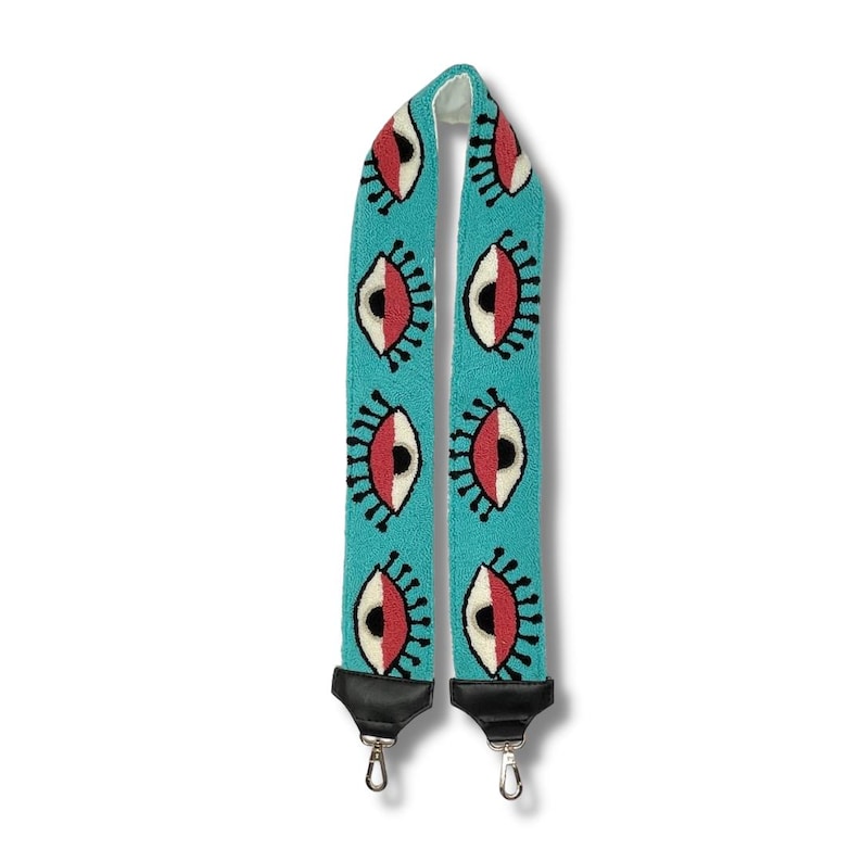 May include: A turquoise blue adjustable strap with a repeating pattern of black outlined eyes with white and red details. The strap has black metal hardware.