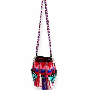 May include: A woven bucket bag featuring a vibrant geometric pattern in red, pink, blue, and turquoise. The bag includes a long, braided chevron-patterned shoulder strap and two decorative tassels hanging from the front with multi-colored threads.