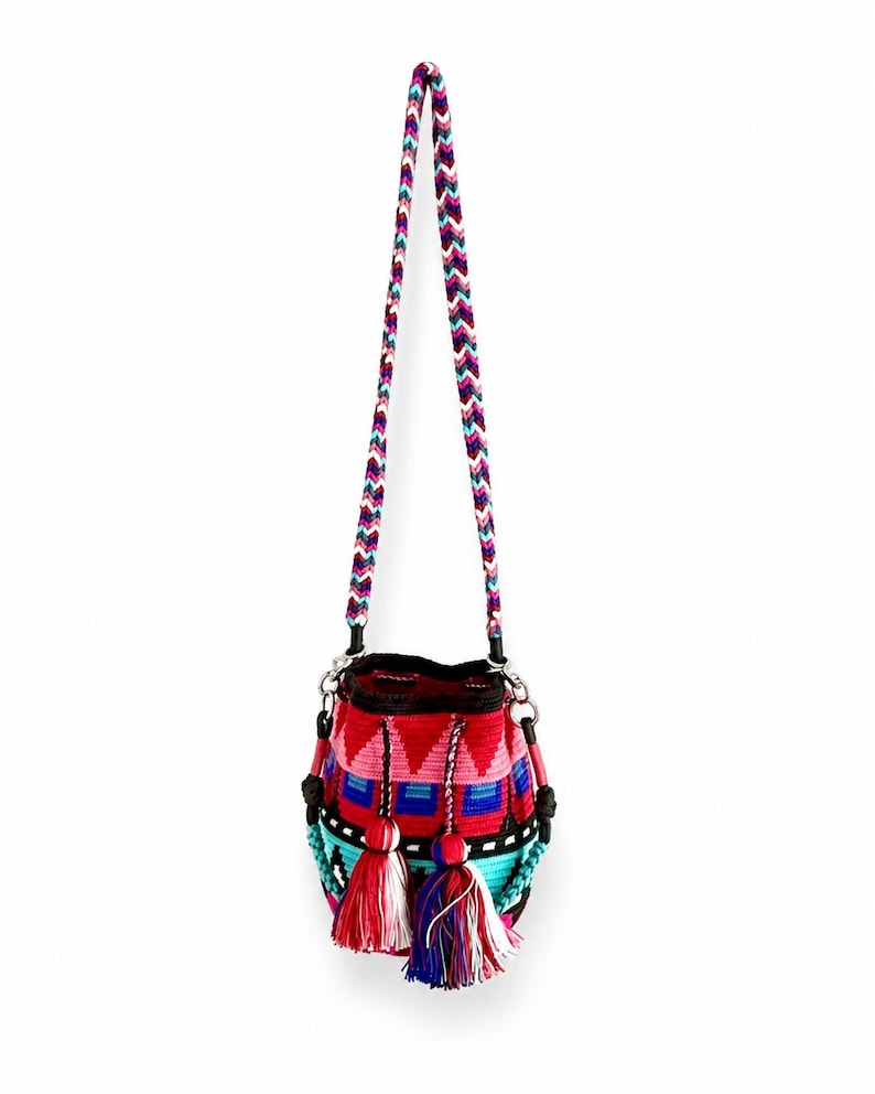 May include: A woven bucket bag featuring a vibrant geometric pattern in red, pink, blue, and turquoise. The bag includes a long, braided chevron-patterned shoulder strap and two decorative tassels hanging from the front with multi-colored threads.