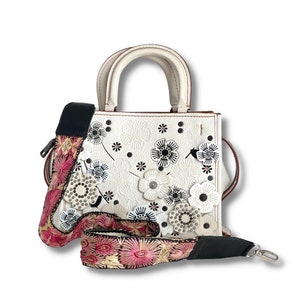 Puede incluir: Bolso de mano de cuero blanco con diseño floral y correa negra y rosa. El bolso tiene un asa superior y una correa de hombro desmontable.