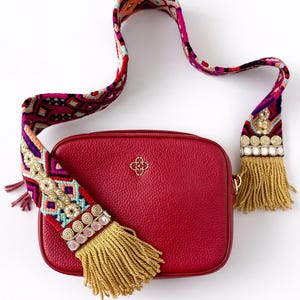 May include: A red leather crossbody bag with a decorative strap. The strap is woven with colorful geometric patterns and embellished with gold-colored tassels and decorative elements. The bag has a gold-colored emblem on the front.